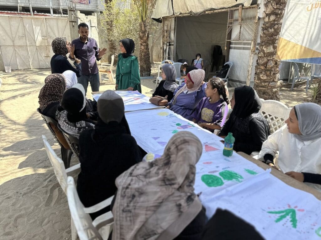 A man speaks before a group of young women wearing hijabs sitting around a table on a dirt floor.