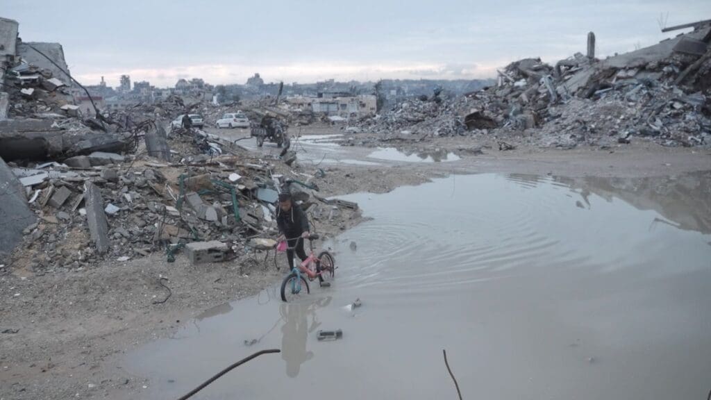 A child pulls a bicycle over still water in the middle of a destroyed city.