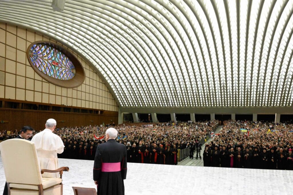 Foto desde la espalda del Papa León, al lado un cardenal. Ambos miran a una multitud en auditorio.