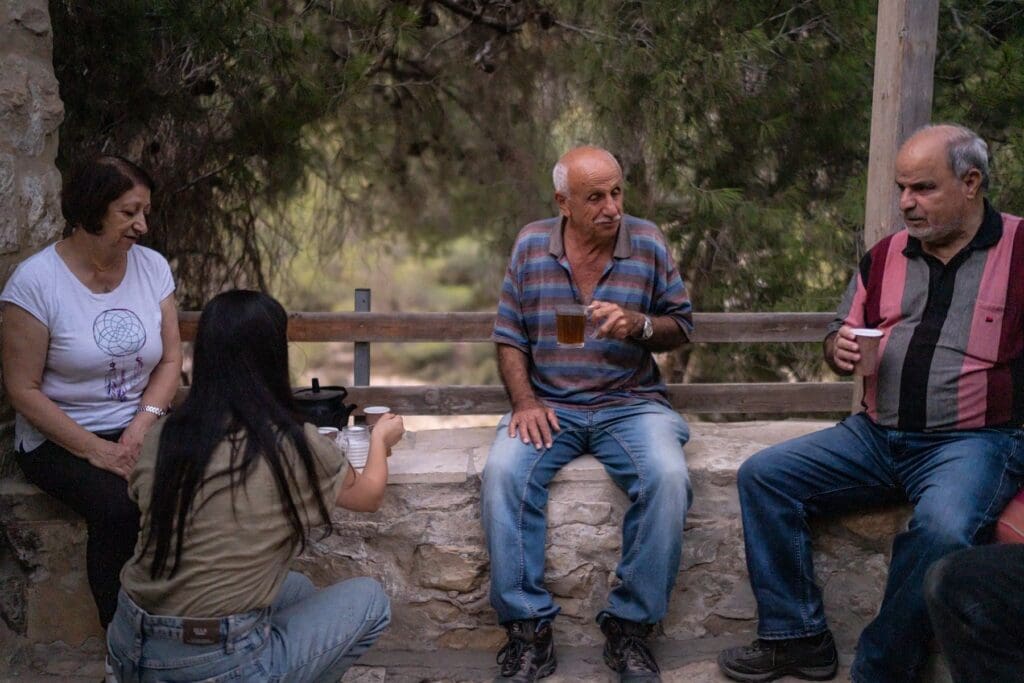 Dos hombres y una mujer están sentados en un muro bajo de contención mientras una joven sirve una taza de té para la mujer.