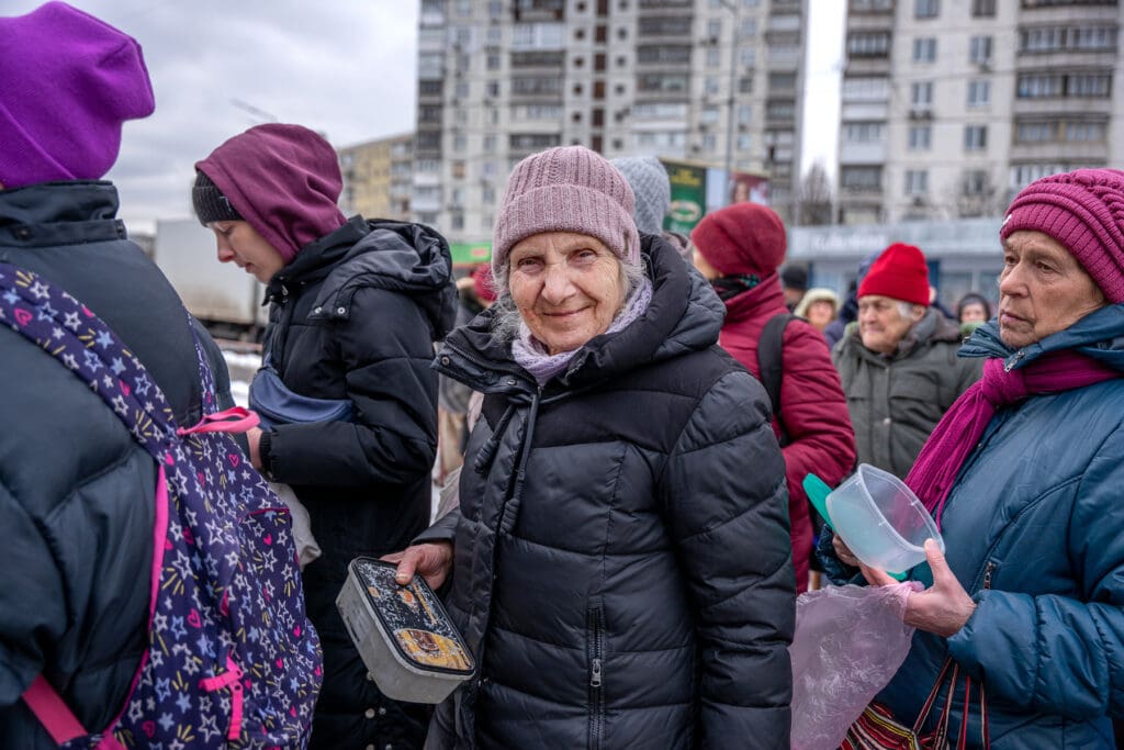 Una anciana con ropa de invierno sonríe mientras espera en fila para recibir comida.