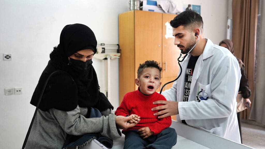 Un médico atiende a un niño que llora, mientras su mamá lo sostiene de una mano.