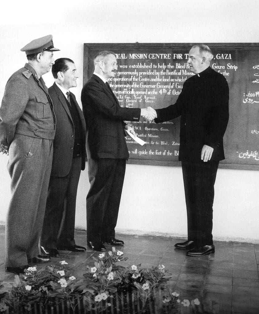 Un sacerdote, de pie frente a una pizarra, estrecha la mano de un hombre, mientras otros dos hombres observan sonriendo.