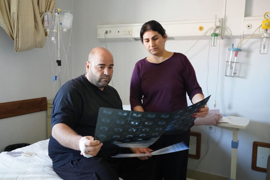 Un hombre sentado en una cama de hospital y una mujer a su lado observan unas radiografías.