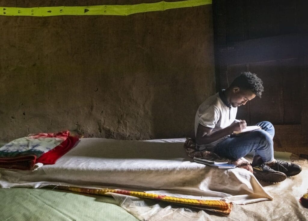 Un ragazzo seduto su un materasso poggiato a terra mentre studia.