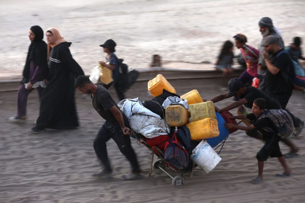 Un uomo tira una carriola piena di oggetti; un bambino lo aiuta spingendo da dietro. Altre persone camminano accanto a loro nella stessa direzione.