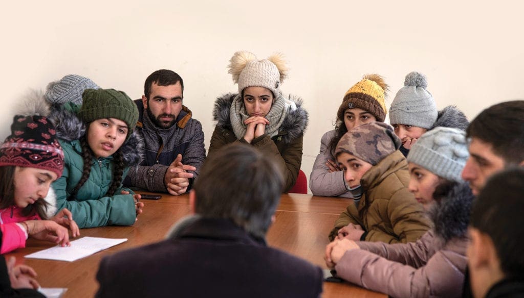 About a dozen youth gather around a table, chatting with a priest in the eastern european nation of armenia.