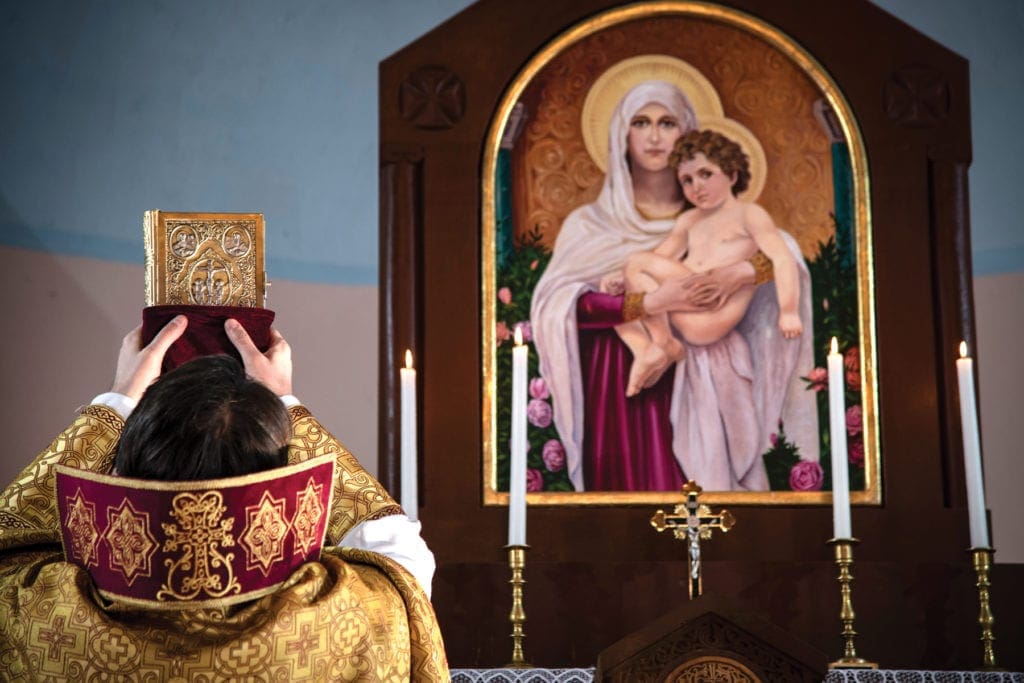 Priest prays at altar with large religious icon in background.