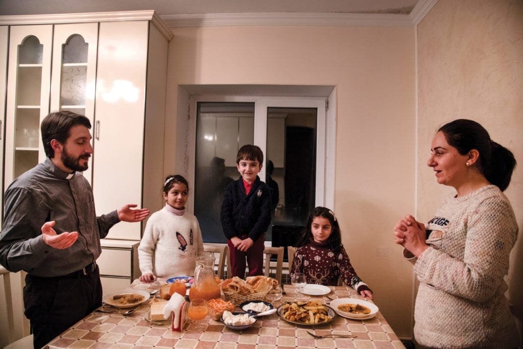 Priest stands opposite wife at dinner table, with three children between, as they all say grace.