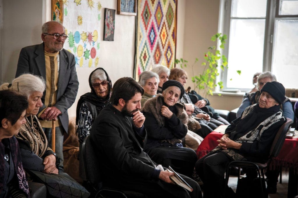 Young priest sits and prays with a room full of seniors citizens.