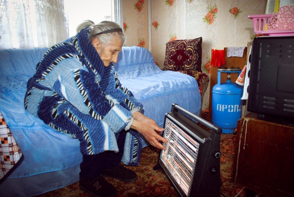 elderly woman in armenia holds her hands in front of a space heater.