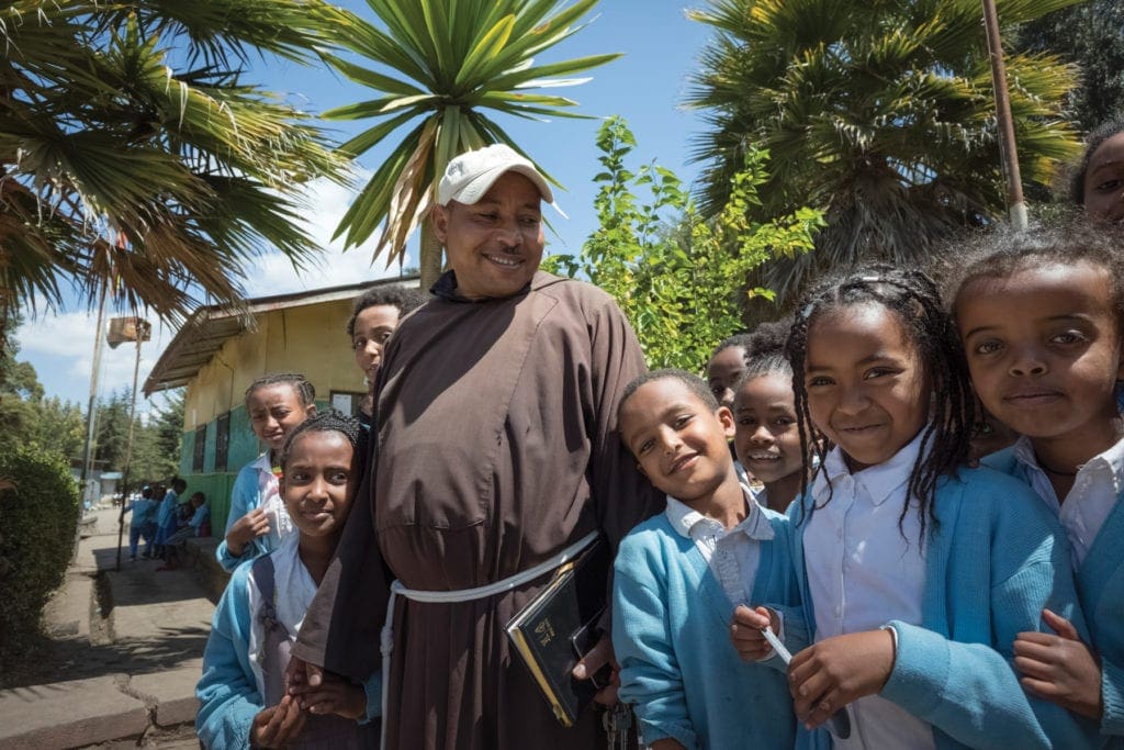 Capuchin priest in baseball cap stands among children outside of school.