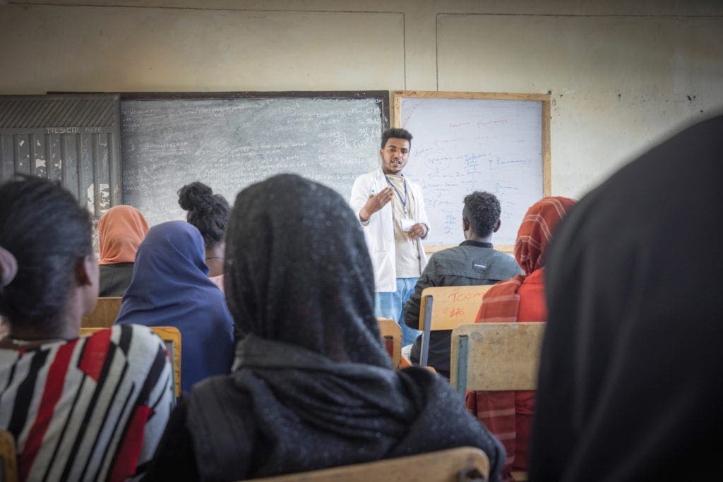 Young man in white coat lectures before a classroom at kidane mehret.