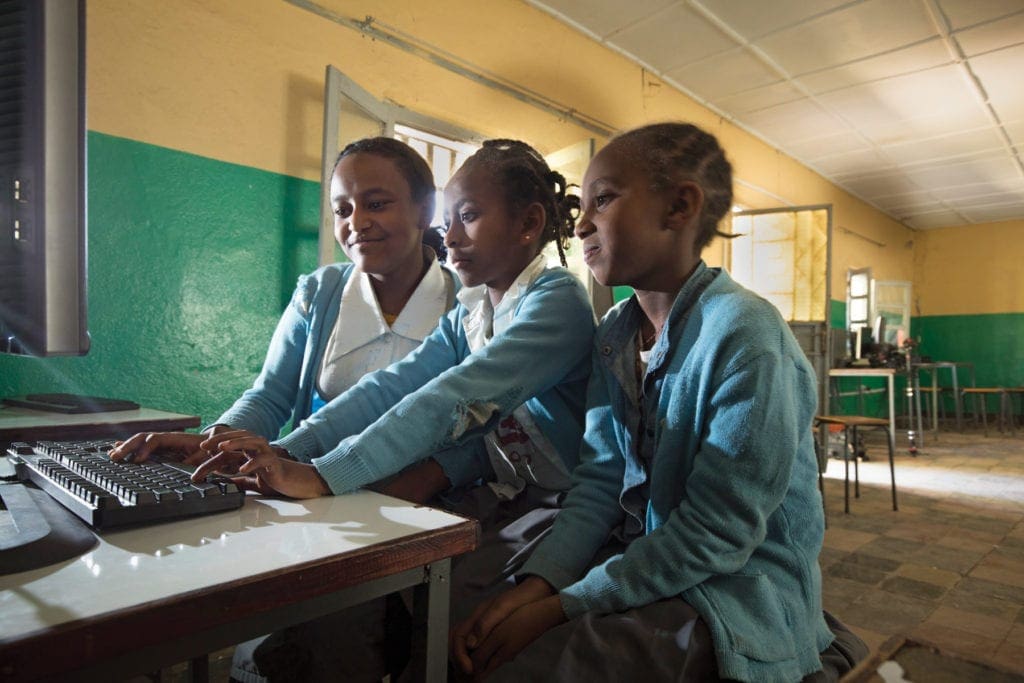 Three youth cluster around one computer, watching it attentively.