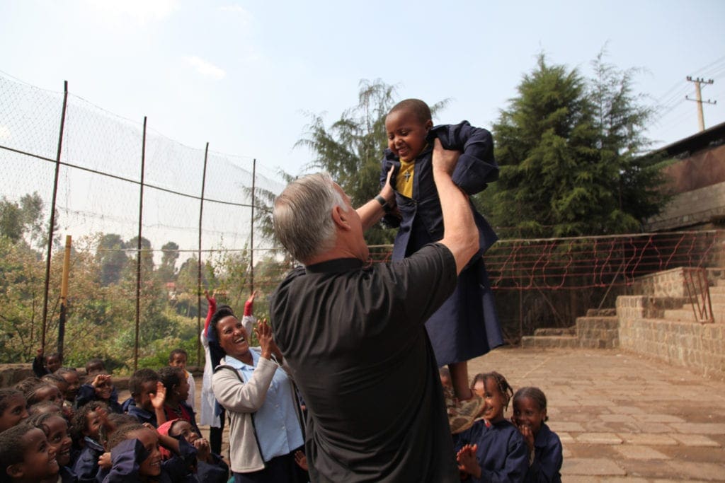 Msgr. holds a grinning child up in the air as his classmates gather around.