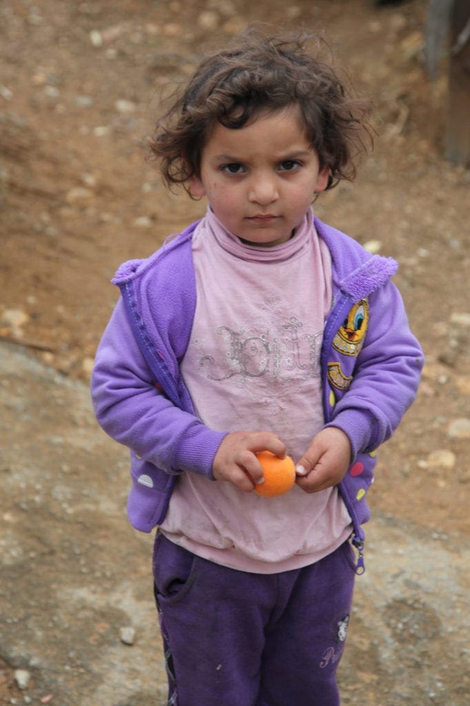 a young girl peers at the camera while holding an orange.