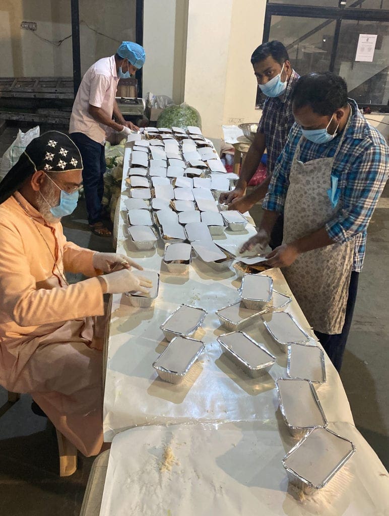 the archbishop and workers in surgical masks gather around a table, sealing containers of cooked food.