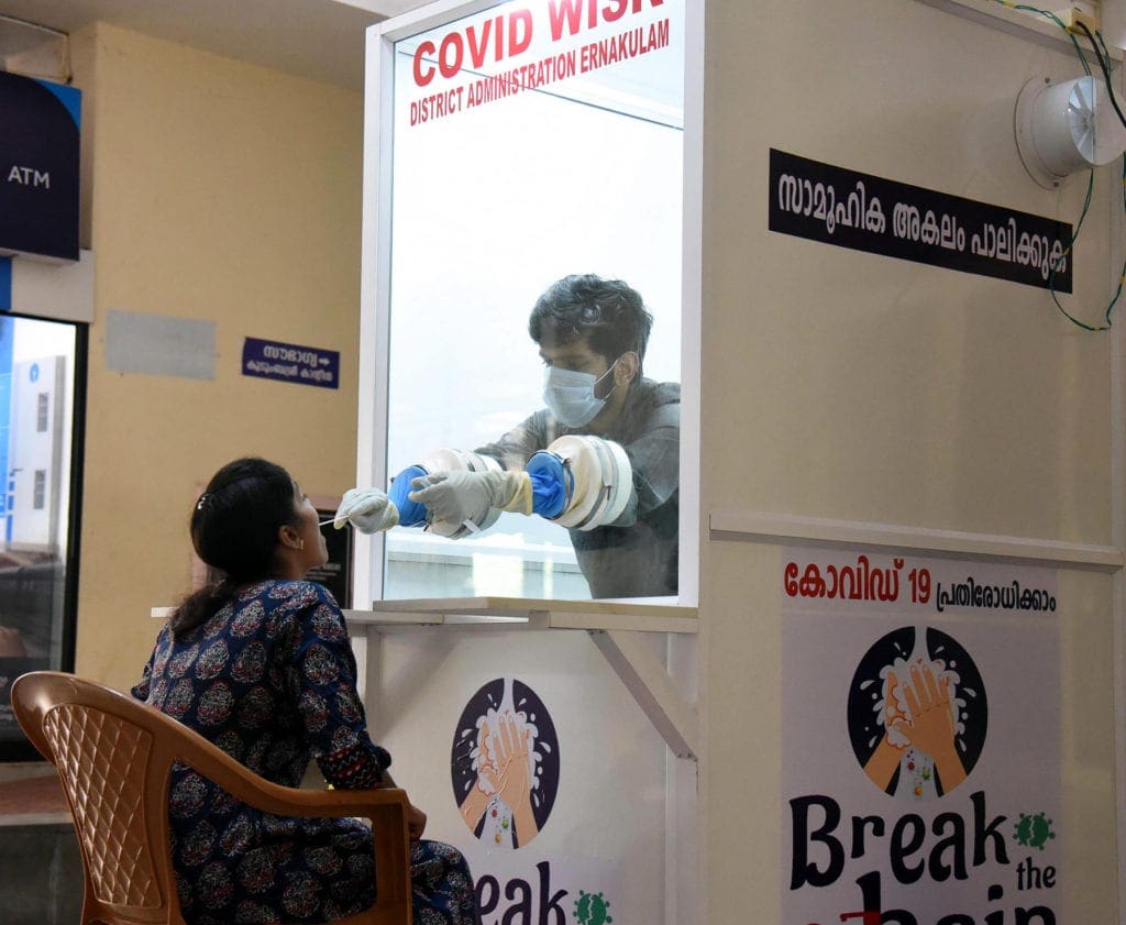 a medical worker reaches through gloves extending from a pane of glass to administer a virus swab to a seated woman..