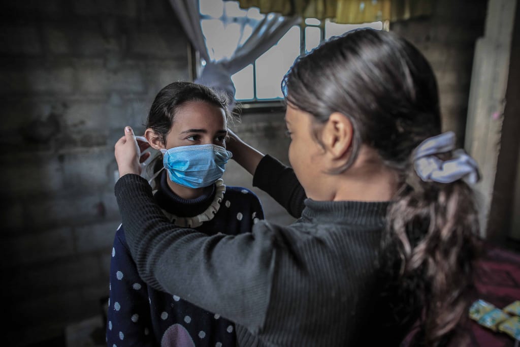 one young woman helps another put on a blue surgical mask.