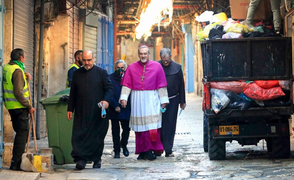 the archbishop and several other men walk past a worker in a safety vest on a narrow street.