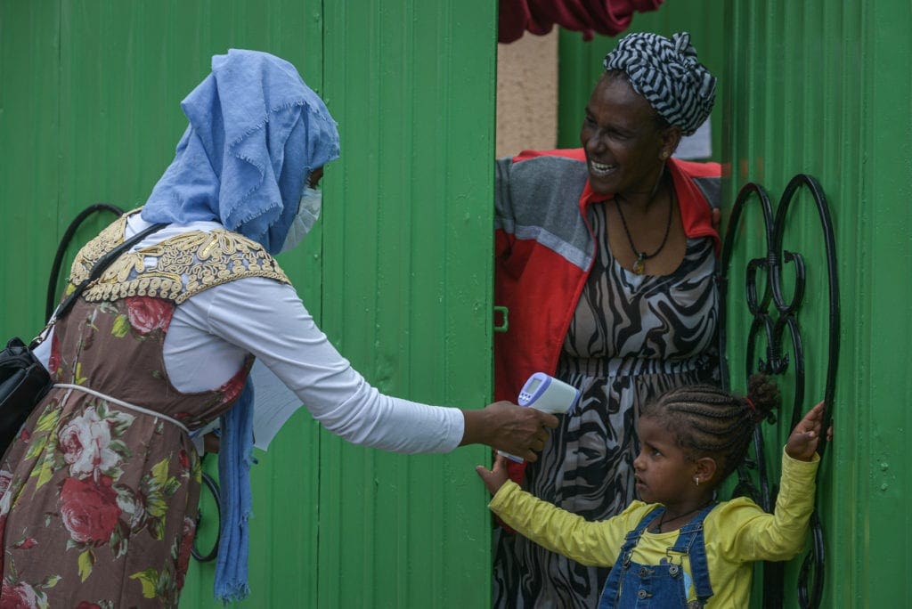 A mother stands at her doorway while a masked woman uses a laser thermometer to take her daughter's temperature from a short distance.