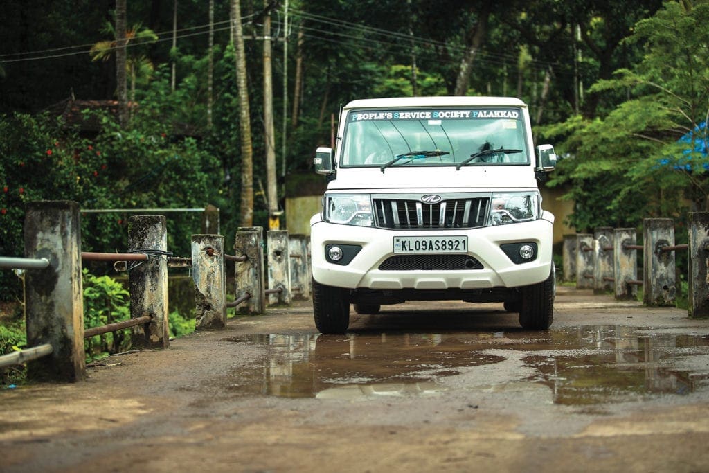 a white truck labeled as belonging to the People's Service Society Palakkad drives through a puddle.