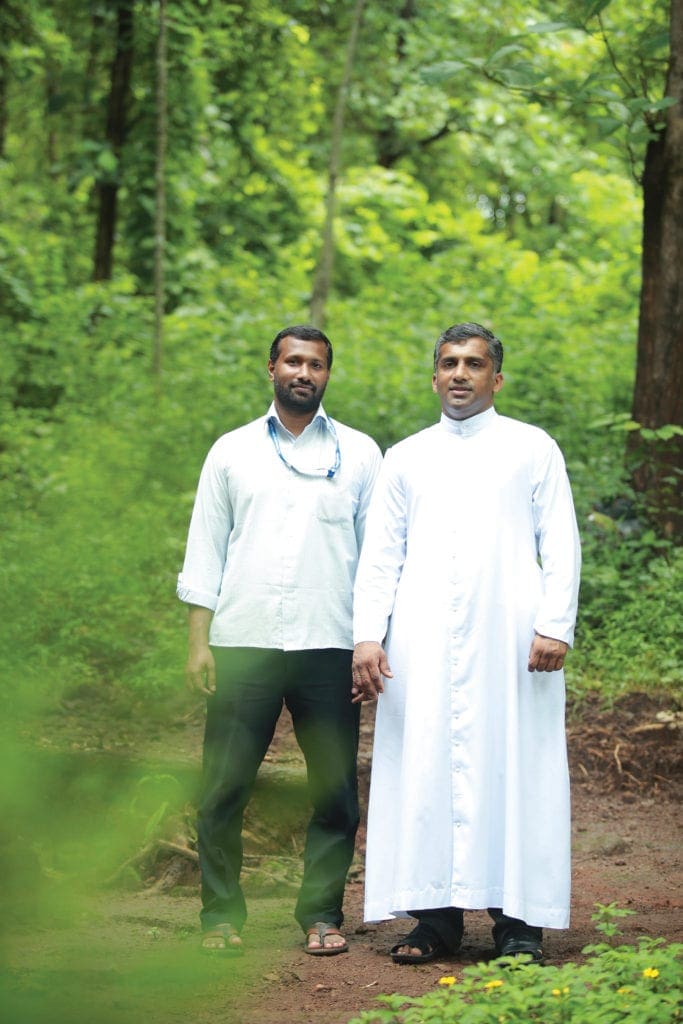 Two priests stand side by side amid bright green woodland flora.