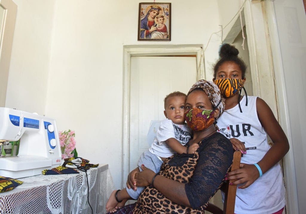 A woman sits before a sewing machine, holding her 1-year-old son while her 10-year-old daughter stands beside.