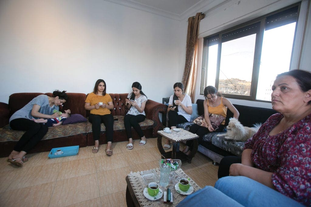 A family sits on a variety of chairs and couches around the perimeter of a small living room.