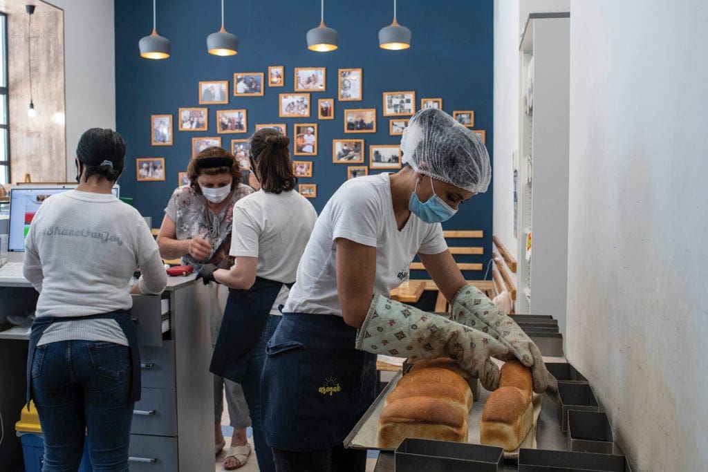 a woman in a hairnet, mask and baking gloves inspects bread fresh from the oven.