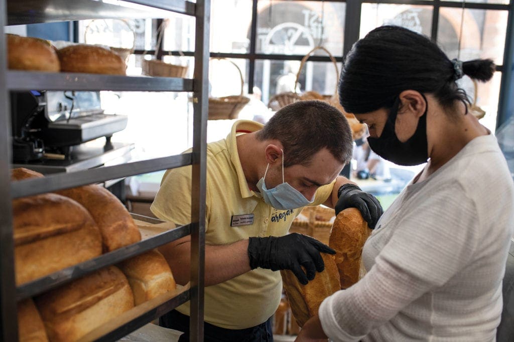 a young man in a mask and gloves takes a pair of loaves off a rack.