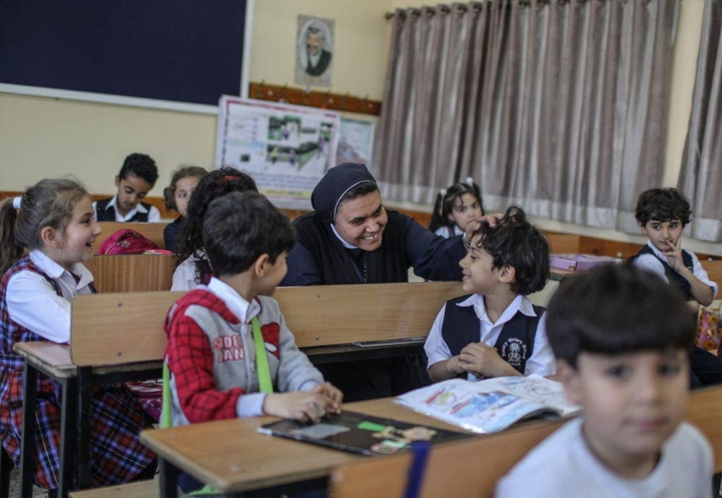 A religious sister sits in the middle of a classroom, tousles the hair of a smiling student in Palestine.