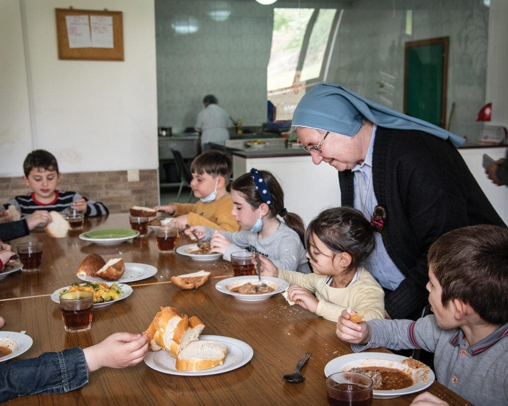 a nun chats with children eating soup at a large table.