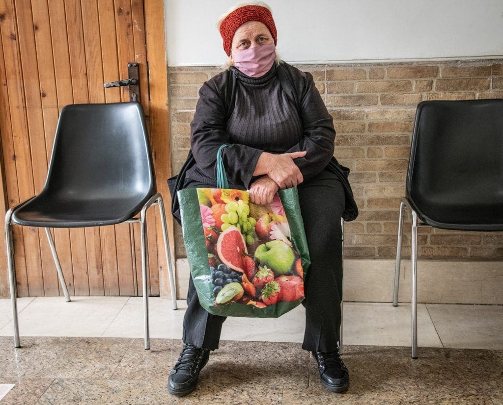 a woman with a mask and a bag is seated near a wall in the caritas soup kitchen.