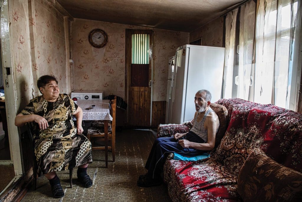 an elderly man and woman sit in a small room in a domik with a couch and kitchen appliances.