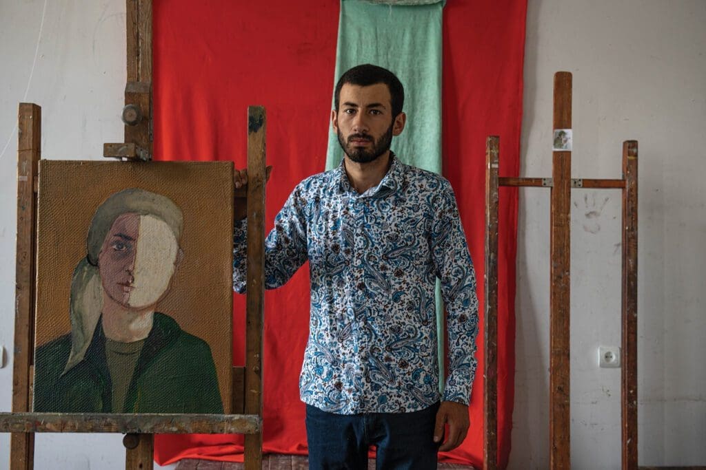 a young man stands beside a painting of a woman with half her face blanked out.