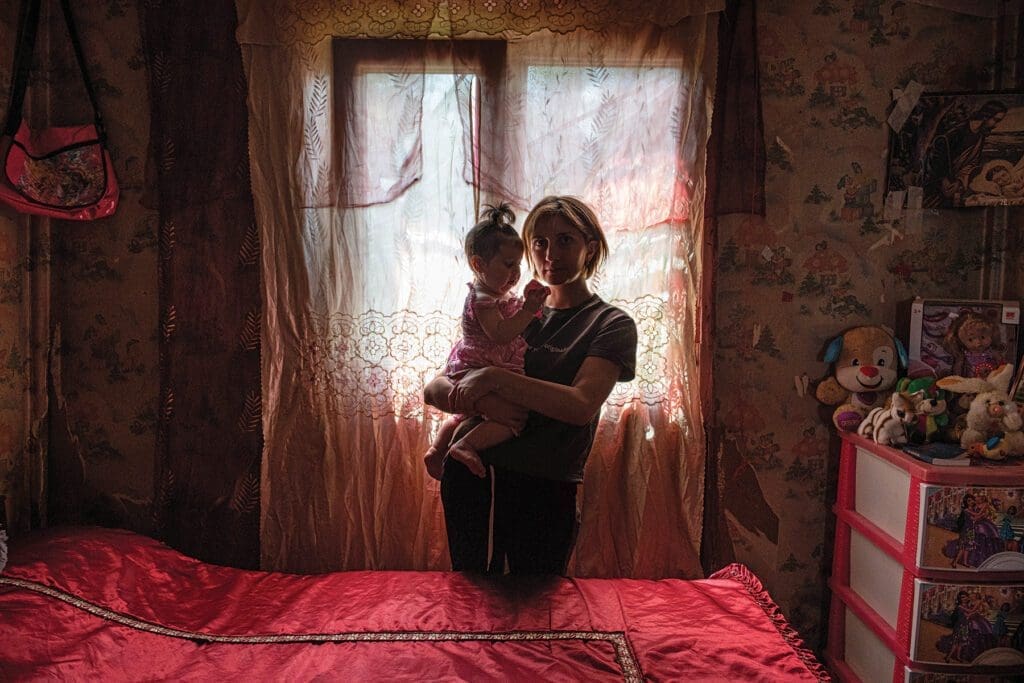 a woman holds her daughter, backlit from a curtained window in a domik.