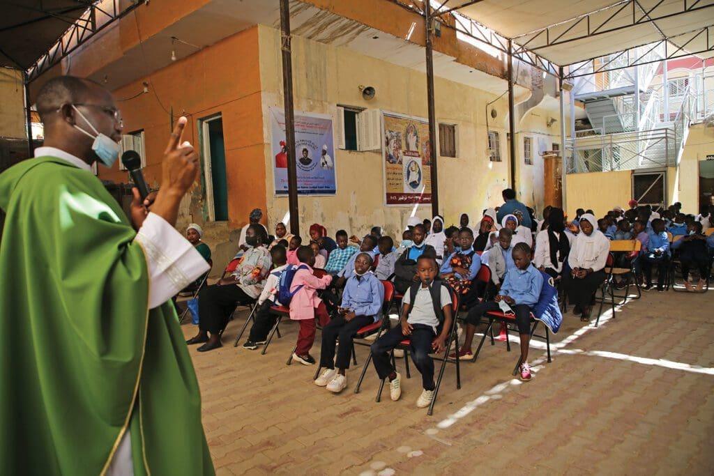 a priest with a microphone addresses a crowd of students on folding chairs.