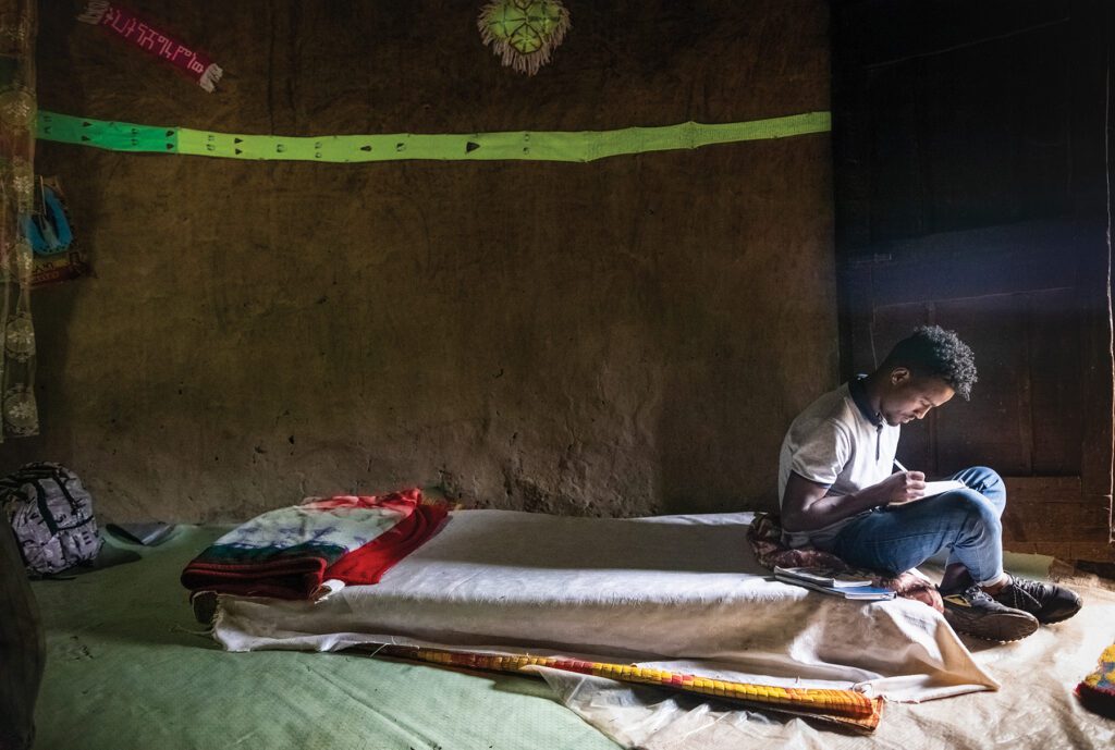 university student sitting on the edge of a bed, writing on papers in his lap.