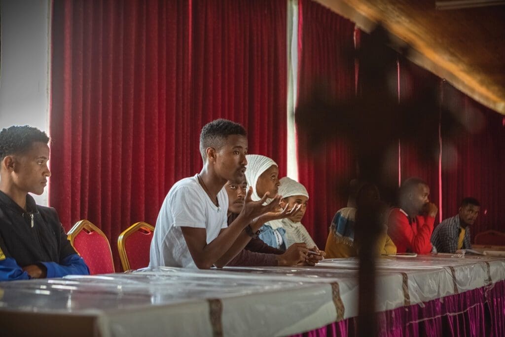 man seated at long table speaks to assembly.