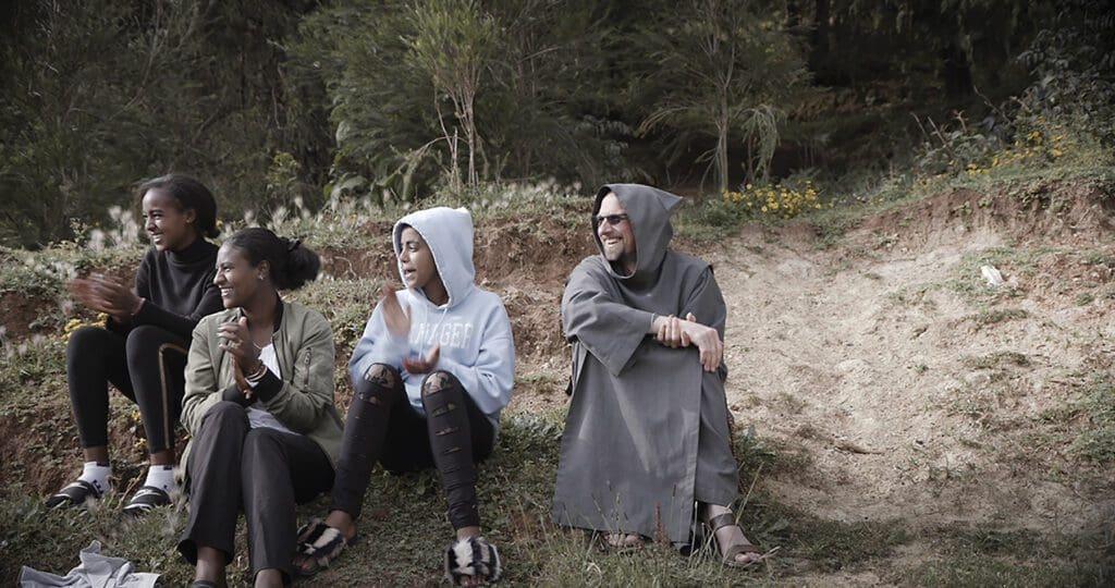 young adults sit on grass joined by a religious man in a hood.