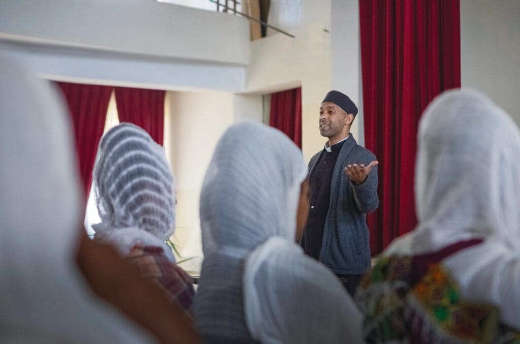 a priest addresses an audience of young women in white headscarves.