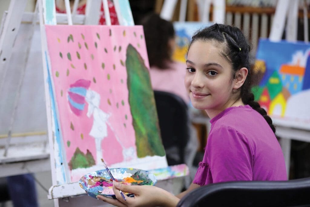 a girl holds a brush and palette beside a painting she is working on.