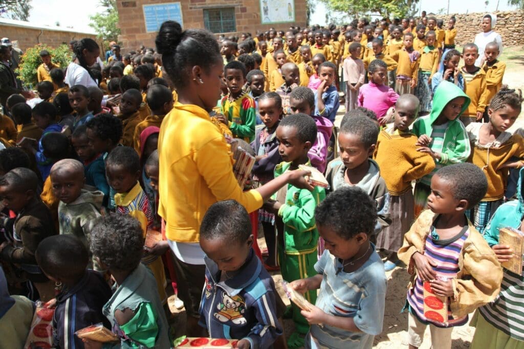 A woman in a yellow shirt distributes food to a large group of children