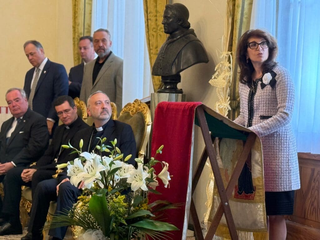 A woman at a podium speaks while men next to her, including a priest listen.