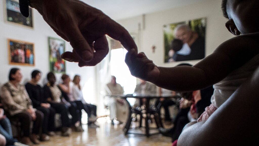 A woman greets an infant, while in the back Pope Francis sits with women.