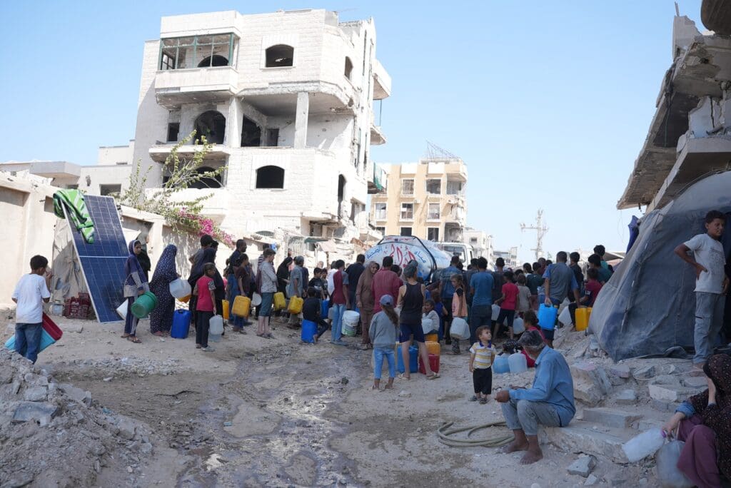 Many people wait in line with containers amid destroyed buildings.