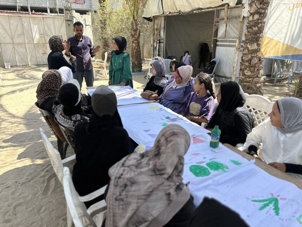 A man speaks before a group of young women wearing hijabs sitting around a table on a dirt floor.