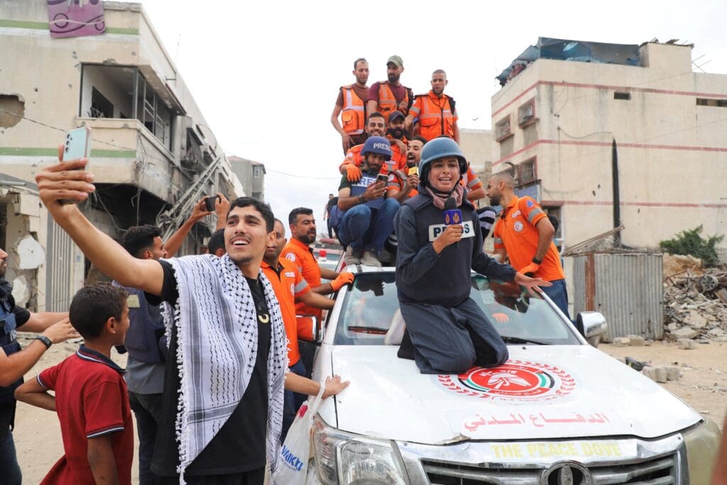 people smiling on top of a car, one of them a woman wearing a helmet and a sweater that reads "press."