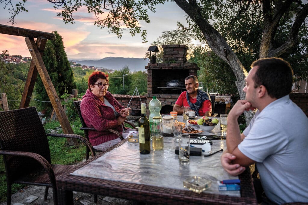 A woman and two men sitting on a porch are having a conversation.